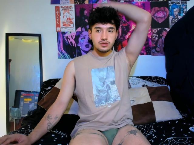 ZorenL, with short curly black hair and a mustache, sits on a bed in a beige tee and green underwear, arm raised, showing tattoos on his arm and thigh.