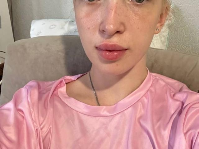 BrookeZapf with shiny pink shirt, shiny lips, freckles, small earrings, and necklace, sitting on beige chair, face slightly up, looking at camera, light skin, subtle makeup.