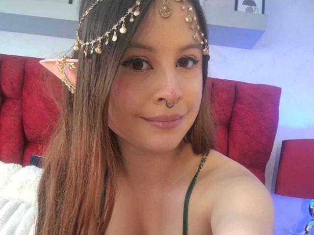 Melani Becker, met bruin haar, een neusring en een gouden hoofdtooi met eraan hangende muntjes, draagt groene bandjes, heeft roze elfenoren en een lichte glimlach, waarbij haar borsten zichtbaar zijn..