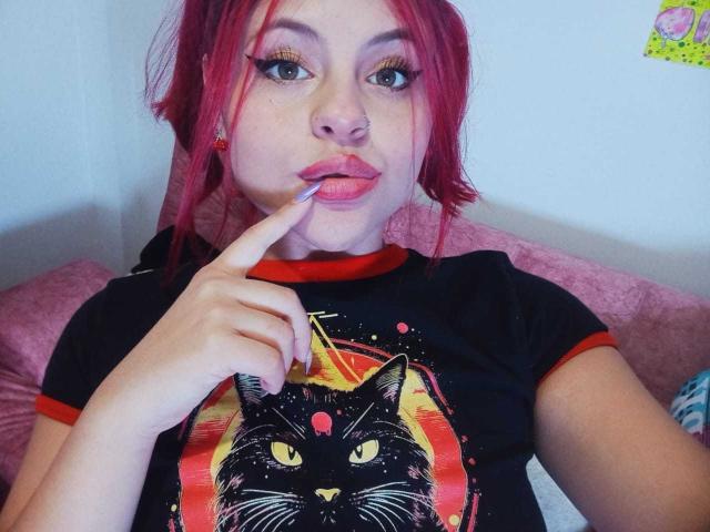 Sophie Hill, con cabello rojo, camiseta negra con estampado de gato, chupándose el dedo, almohada rosa de fondo, cejas rojas, ojos verdes, piercing en la nariz y aretes rojos..