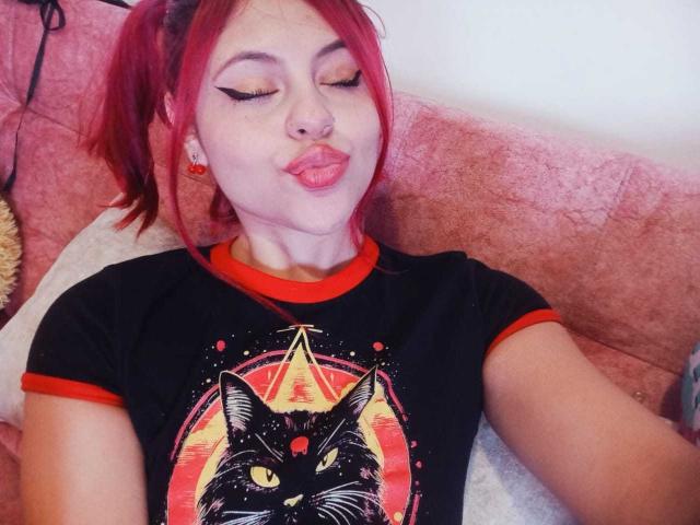 SophieHill with red pigtails, closed eyes, and puckered lips, wearing a black shirt with a cat graphic and red trim, sitting on a pink couch.