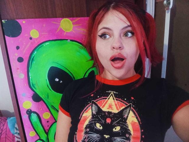 SophieHill with red pigtails, surprised expression, black tee with cat and UFO, green alien painting in background, wide-eyed, pink lips, nose ring, and red eyeliner.