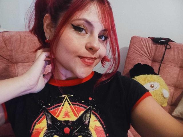 SophieHill with red hair, black shirt, cat graphic, and red trim, smiling, hand in hair, white wall, pink couch, yellow stuffed toy, and black headphones in background.