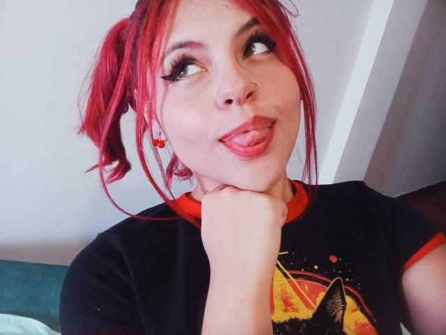 Sophie Hill, avec ses couettes rousses, son t-shirt noir avec des détails rouges, la langue tirée, la tête appuyée sur sa main, un anneau au nez et de longs cils, regardant vers le haut avec une expression espiègle..