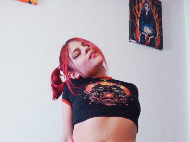 Sophie Hill, avec des couettes rousses, les yeux fermés et les lèvres légèrement entrouvertes, portant un t-shirt noir court avec un motif de crâne, dévoilant ses seins, dans une pièce aux murs blancs..