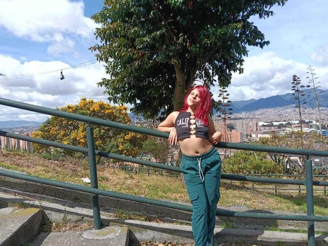 SophieHill with red hair, green pants, and a lifted gray top exposing her tits, leaning on a green railing, standing on concrete steps, sunny day, city in background.