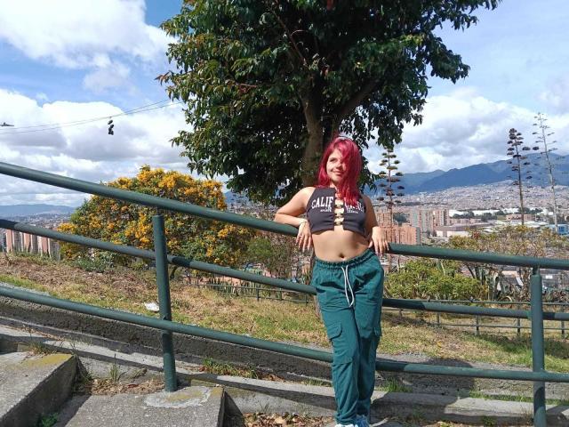 SophieHill with red hair, small tits, green pants, black crop top, leaning on green rail, grassy hill, tree, city in background, clear sky, mountains, gondola, casual, sunny.