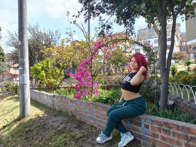 SophieHill with red hair, black crop top, green pants, and white sneakers, sitting on a brick wall, leaning on a tree, with purple flowers and greenery in the background.