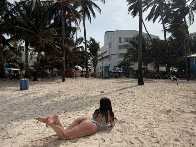 AnyaJay lies on her stomach on a sandy beach, wearing a green thong bikini, with her bare ass and feet up, palm trees and a white building in the background.