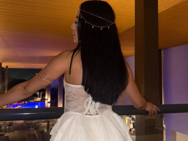 AnyaJay with long black hair, wearing a white lace dress, arm bracelet, and head chain, standing with back to camera, hands on railing, in dimly lit room.