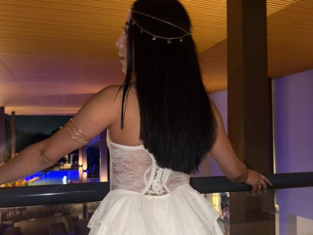 AnyaJay with long black hair, wearing a white lace-up dress and gold headpiece, standing with arms on a railing, showing her back and a gold arm bracelet.