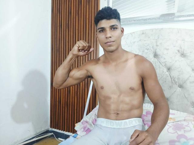SantiaGodiazz, sem camisa, musculoso, pele bronzeada, flexionando o braço direito, cabelo preto encaracolado, calças brancas HANDBRAVERS, sentado na cama, expressão confiante, iluminação suave, quarto com painel de madeira e parede branca..