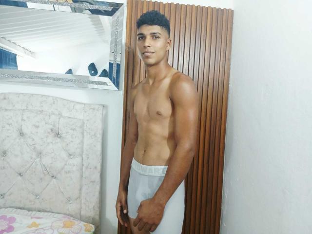 SantiaGodiazz, shirtless, standing, wearing white tighty-whities, muscular, tan skin, short curly black hair, small brown nipples, wood panel door, white wall, mirror, tufted headboard, floral bedsheet.
