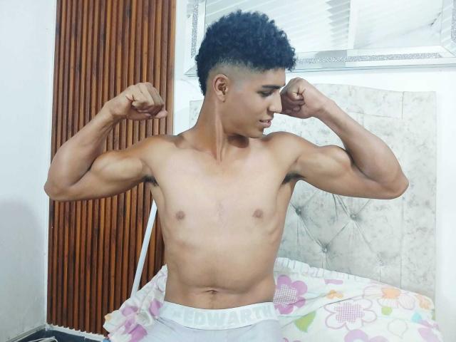 SantiaGodiazz is a muscular, shirtless, tan-skinned guy with curly black hair, flexing his biceps, wearing white underwear, standing in a room with a floral bedsheet.