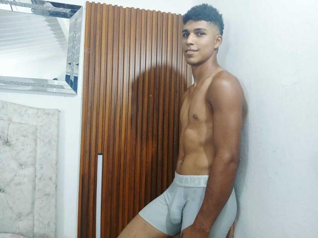 SantiaGodiazz, sem camisa, com um corpo magro e tonificado, vestindo roupa íntima cinza justa, em pé contra uma parede de madeira, cabelo curto e encaracolado, expressão séria, pele marrom clara..
