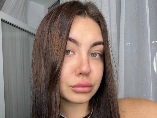 Ines Gueningsman tem cabelo castanho liso, olhos verdes e lábios rosados com uma expressão neutra. Ela está usando um colar preto. Seu nariz é pequeno e sua pele é clara..