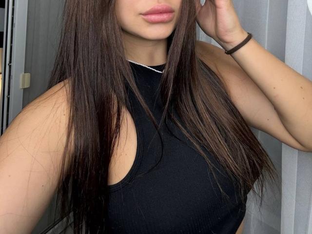 InesGueningsman with long brown hair, wearing a black sleeveless top, has pink lips, a black bracelet on her right wrist, and her right hand touching her hair.