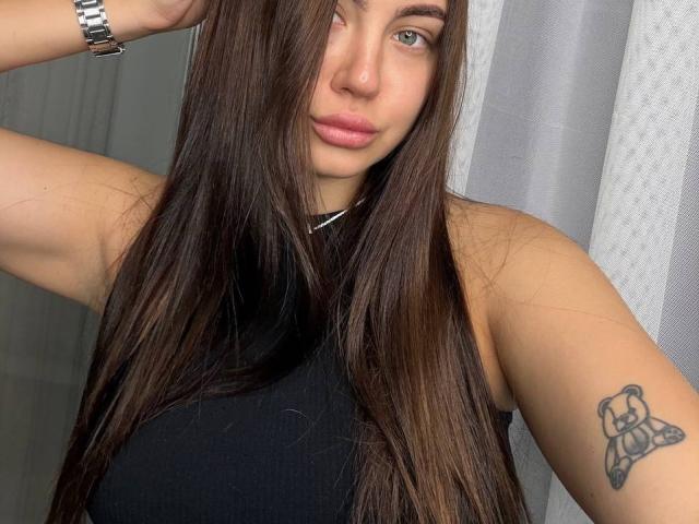 Ines Gueningsman tem cabelos castanhos longos, olhos azuis e uma blusa preta sem mangas, que mostra uma tatuagem de um urso em seu braço direito, e está usando um relógio de pulso prateado..