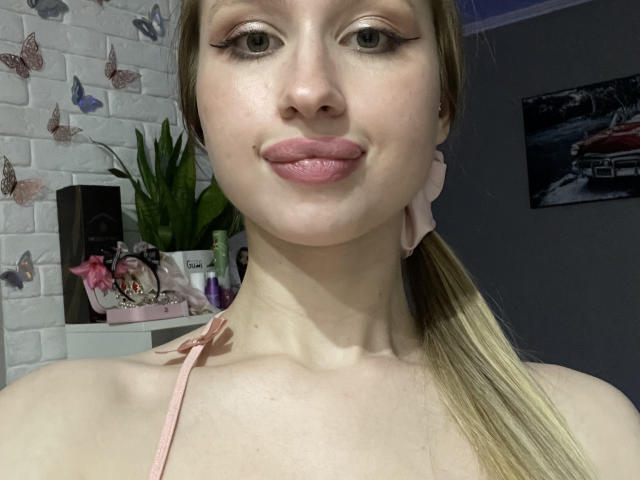 Leonida Ferdolage a la peau pâle, une queue de cheval blonde, du rouge à lèvres rose et du crayon pour les yeux noir. Elle porte un élastique rose autour du cou et un haut rose à bretelles fines..
