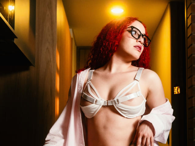 LilJane with curly red hair, black glasses, white strappy bra exposing her tits, and pink shirt off, looking up in a yellow-lit hallway.
