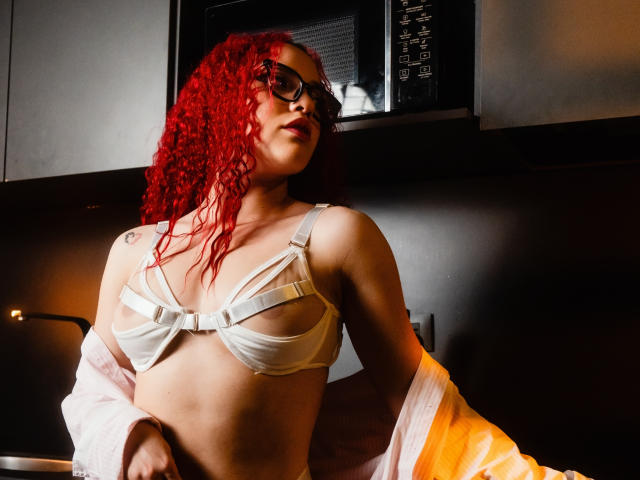 LilJane, con cabello rizado de color rojo brillante, usando gafas, un sostén blanco con tirantes y una camisa rosa que se desliza del hombro, mostrando sus pequeños senos y un tatuaje en su brazo izquierdo..