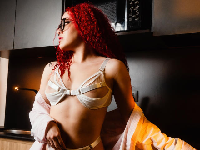 LilJane, con i capelli rossi e ricci, indossa occhiali neri, un reggiseno bianco e una maglia trasparente che lascia intravedere i suoi seni, è in piedi in una cucina e guarda di lato. Ha un tatuaggio..