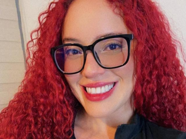 LilJane, con i capelli rossi e ricci, gli occhiali neri e il rossetto rosso, sorride alla fotocamera, indossando una giacca verde scuro, ha un viso carino e attraente..