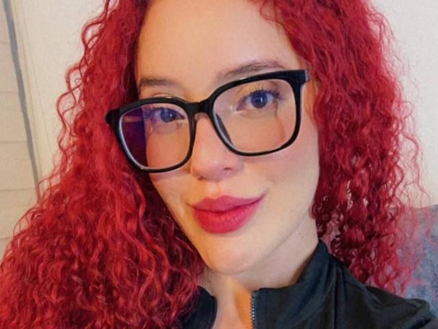 LilJane with curly red hair, black glasses, and pink lipstick, wearing a black zip-up shirt, smiling at the camera. Her blue eyes are slightly squinted.