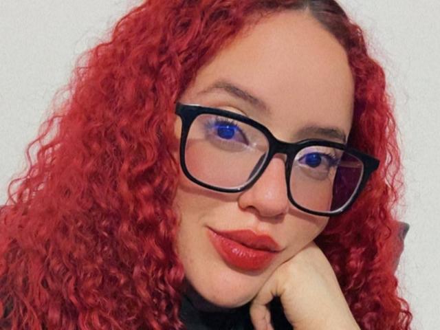 LilJane has red curly hair, blue eyes, black glasses, and red lipstick. She's resting her chin on her hand with a slight smile. Boobs not visible.