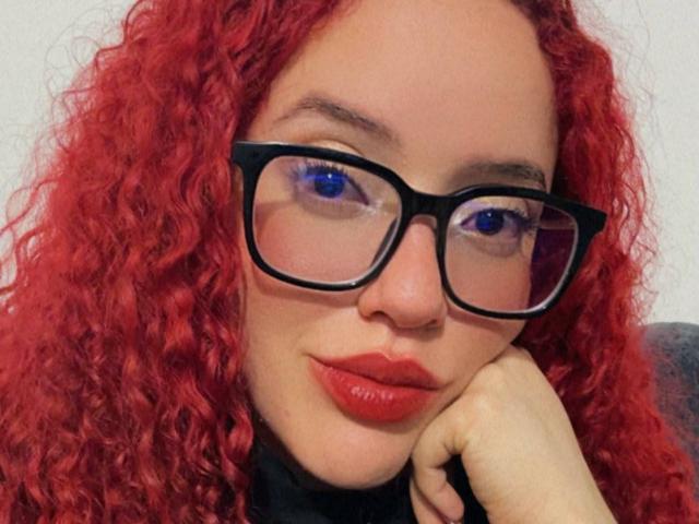 LilJane, con i capelli rossi ricci, gli occhiali neri, gli occhi azzurri e le labbra rosse, appoggia la testa sulla mano, mostrando il suo viso carino e i suoi occhiali sexy..