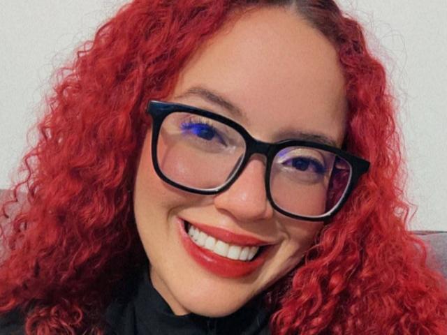 LilJane with curly red hair, black glasses, and red lipstick, smiling with bright blue eye makeup, showing her white teeth. Her tits are not visible.