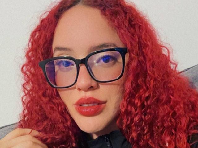 LilJane, con cabello rojo rizado, gafas negras y lápiz labial rojo, mostrando su linda cara y labios ligeramente entreabiertos, vestida con una blusa negra..