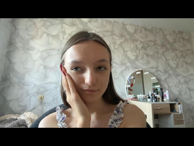 LachelleTrivino has a small nose, long brown hair, and a hand on her cheek. She's wearing a floral tank top, showing her tits, in a floral wallpapered room.