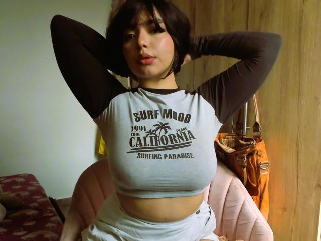 AnnieUwU with huge tits, wearing a grey and brown raglan shirt, hands behind head, sitting on a pink chair, looking at camera, brown hair, wooden wall in background.