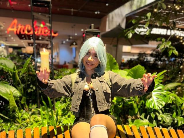 AnnieUwU with teal hair, smiling, arms out, wearing a green jacket, black top, and shorts, sitting on a wooden bench, holding a yellow drink, lush green plants around.