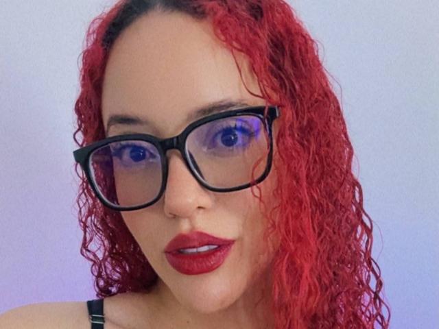 LilJane mit lockigem, rotem Haar, die eine schwarze Brille und roten Lippenstift trägt, hat blaue Augen und ein leichtes Lächeln und blickt direkt in die Kamera. Ihre Brüste sind teilweise durch den schwarzen Träger zu sehen..