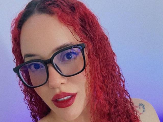 LilJane with red curly hair, black glasses, and red lipstick, looking at the camera with blue eyes, showing a small tattoo on her right shoulder.