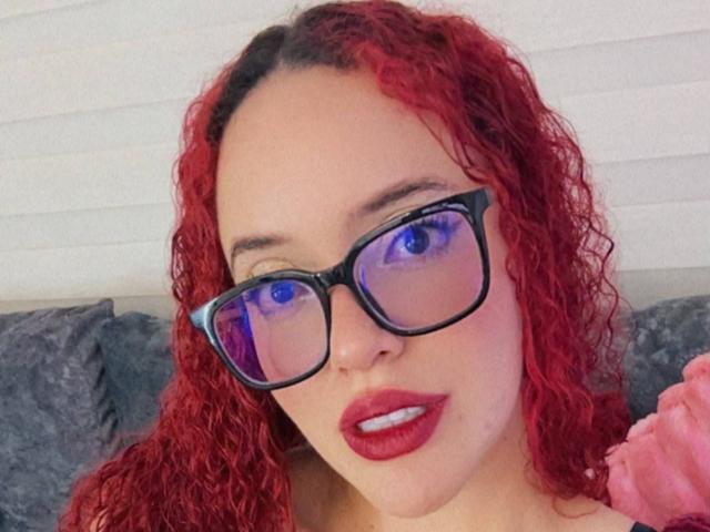 LilJane tiene el cabello rojo y rizado, gafas negras con reflejos morados y lápiz labial rojo. Está mirando a la cámara con una ligera sonrisa, mostrando sus dientes blancos..