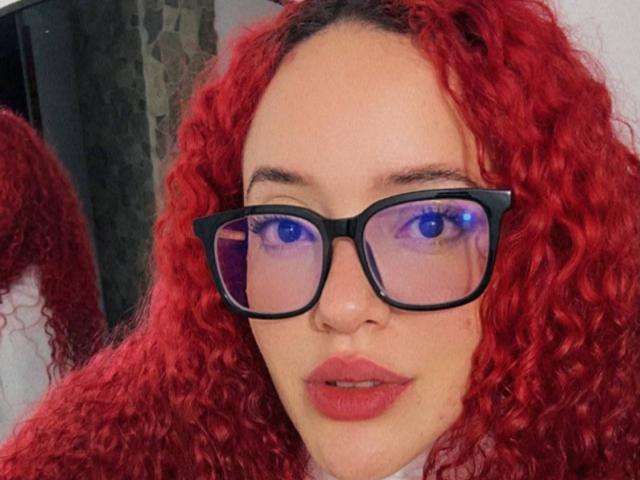 LilJane, com cabelos ruivos encaracolados e vibrantes, óculos de aro preto, olhos azuis brilhantes e batom rosa. Ela está olhando diretamente para a câmera com a boca ligeiramente aberta, de forma sedutora..