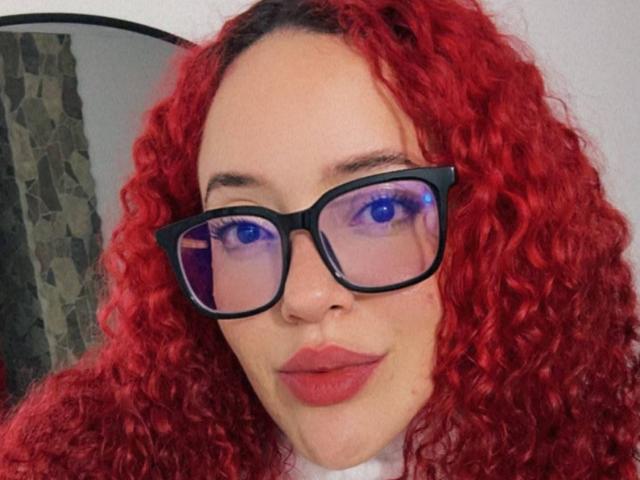 LilJane hat lockiges, rotes Haar, eine schwarze Brille mit blauen Reflexionen und pinken Lippenstift. Sie blickt mit einem leichten Lächeln in die Kamera. Ihre Brüste sind nicht sichtbar..