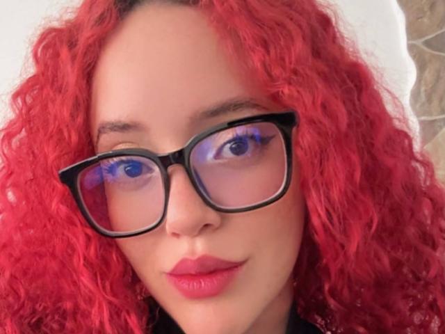 LilJane with curly red hair, black glasses, and pink lipstick, has blue eyes and a slight smile. Her tits aren't visible, just her face.