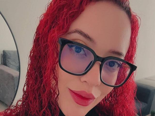 LilJane with red curly hair, blue eyes, and black glasses, wearing red lipstick, smiling at the camera, showing her nose and lips, wearing a black top.