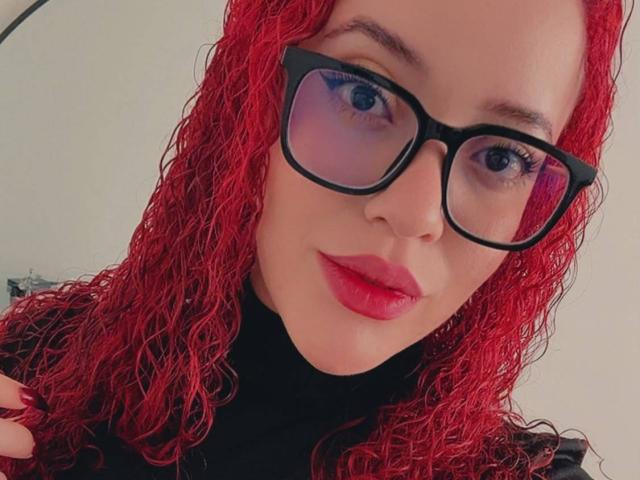 LilJane with curly red hair, black glasses, and red lipstick, wearing a black turtleneck, showing a confident face, her hand near her head.