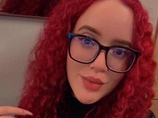 LilJane, con i capelli rossi ricci, indossa occhiali neri e un maglione nero a collo alto, e sorride dolcemente alla fotocamera. I suoi occhi azzurri sono visibili attraverso il vetro..