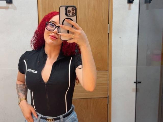 LilJane, com cabelo ruivo encaracolado, óculos, camisa preta com zíper que mostra seus seios, braço tatuado e jeans azuis, tirando uma selfie no espelho com um iPhone dourado..
