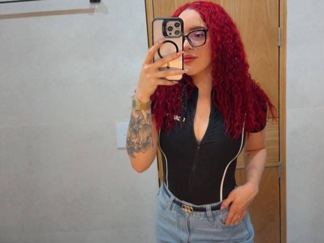 LilJane with red curly hair, glasses, black zip-up top showing her tits, blue jeans, tattooed arm, holding phone, mirror selfie, wooden door in background.
