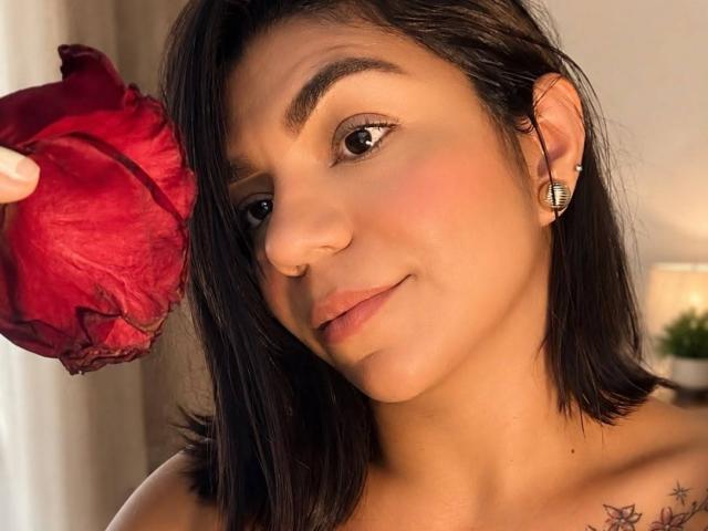 ValeryPierce with short black hair, holding a red rose to her face, has a nose ring, small earrings, and a tattoo on her right shoulder, smiling softly.