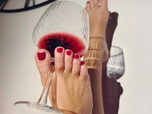 MindyLove's red-painted toes holding a wine glass with pink rosé, her legs crossed, a clear glass in the background, shadows on the white floor.