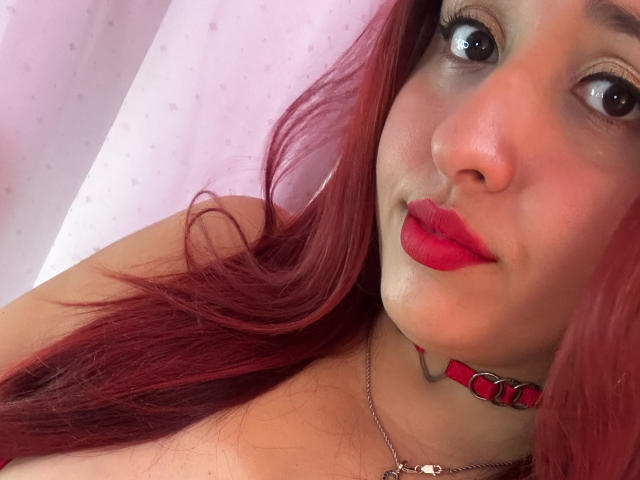 Cassie Miller hat roten Lippenstift, rotes Haar und eine rote Halskette mit Metallringen. Sie blickt mit großen Augen nach oben, und ihre Nase ist leicht nach oben gebogen. Ihre Brüste sind nicht sichtbar..