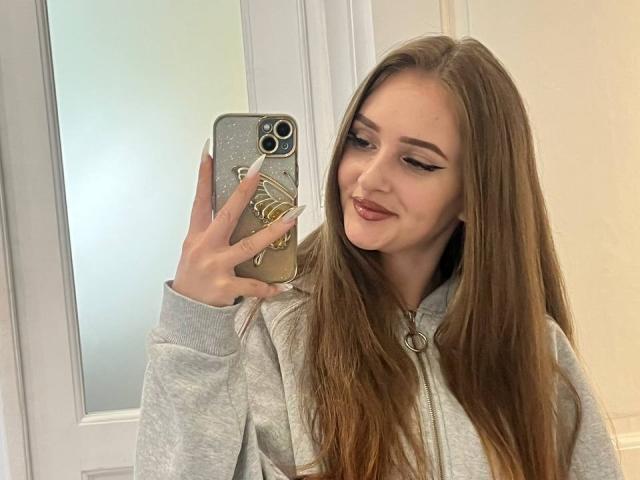 PamelaXu with long brown hair, wearing a grey hoodie, holding a phone with a gold case, smiling, winged eyeliner, and pink lipstick, taking a selfie.